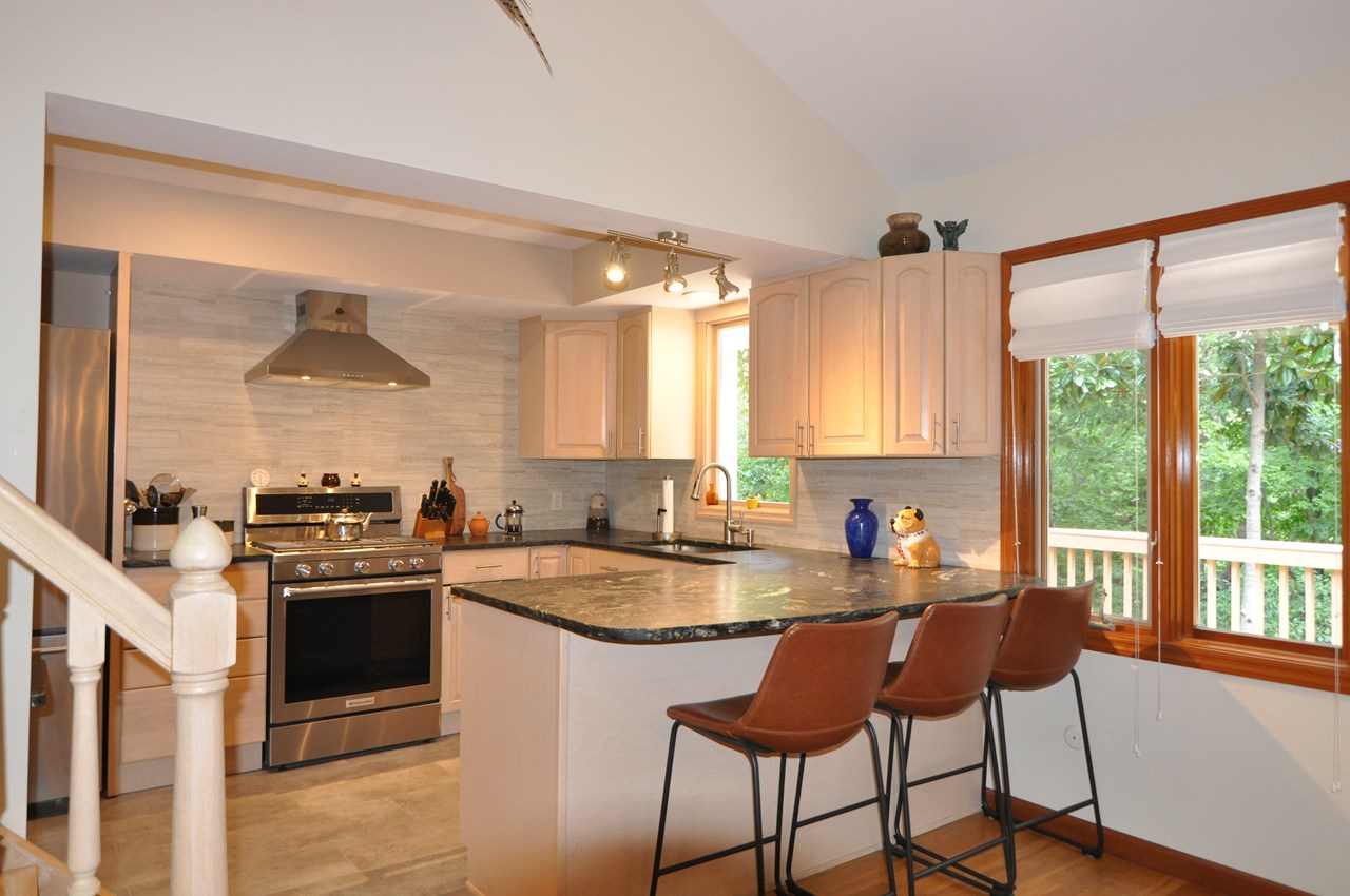 kitchen the kitchen is off the dining area and irregular in size, but approximately 9 x 15. the floor and backsplashes are tile and the counters are honed granite. there is a breakfast bar and the appliances are all newer. there is a built in hood over the gas range and a farm style sink. there is a built in pantry cabinet and there is a shelf for a free standing microwave. the windows look out to the back deck.