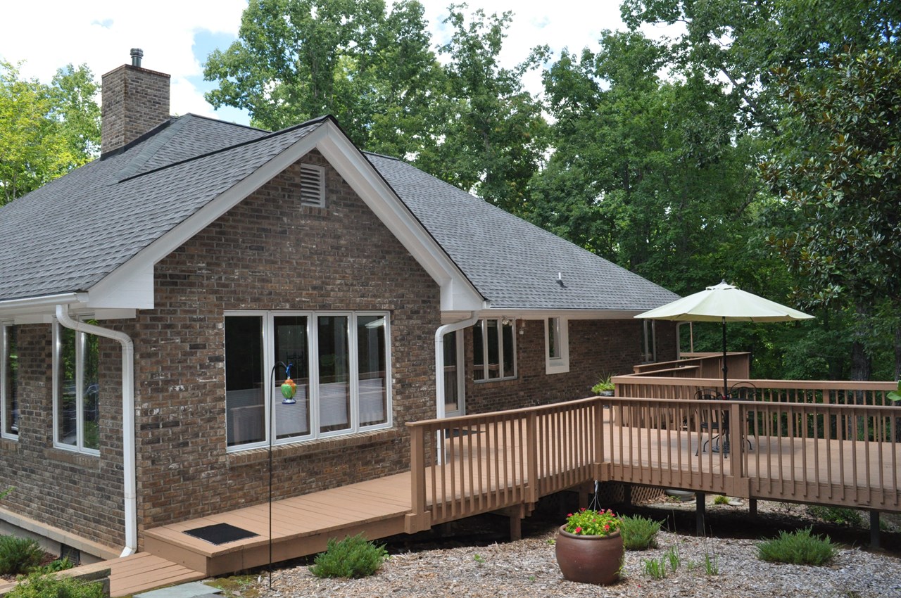 back of the home a stone walk takes you around the right side of the home to the deck. the sitting room off the primary bedroom is shown to the left and it has direct access to the deck. the deck wraps around to the back left side of the home with an entrance to the back guest bedroom as well.